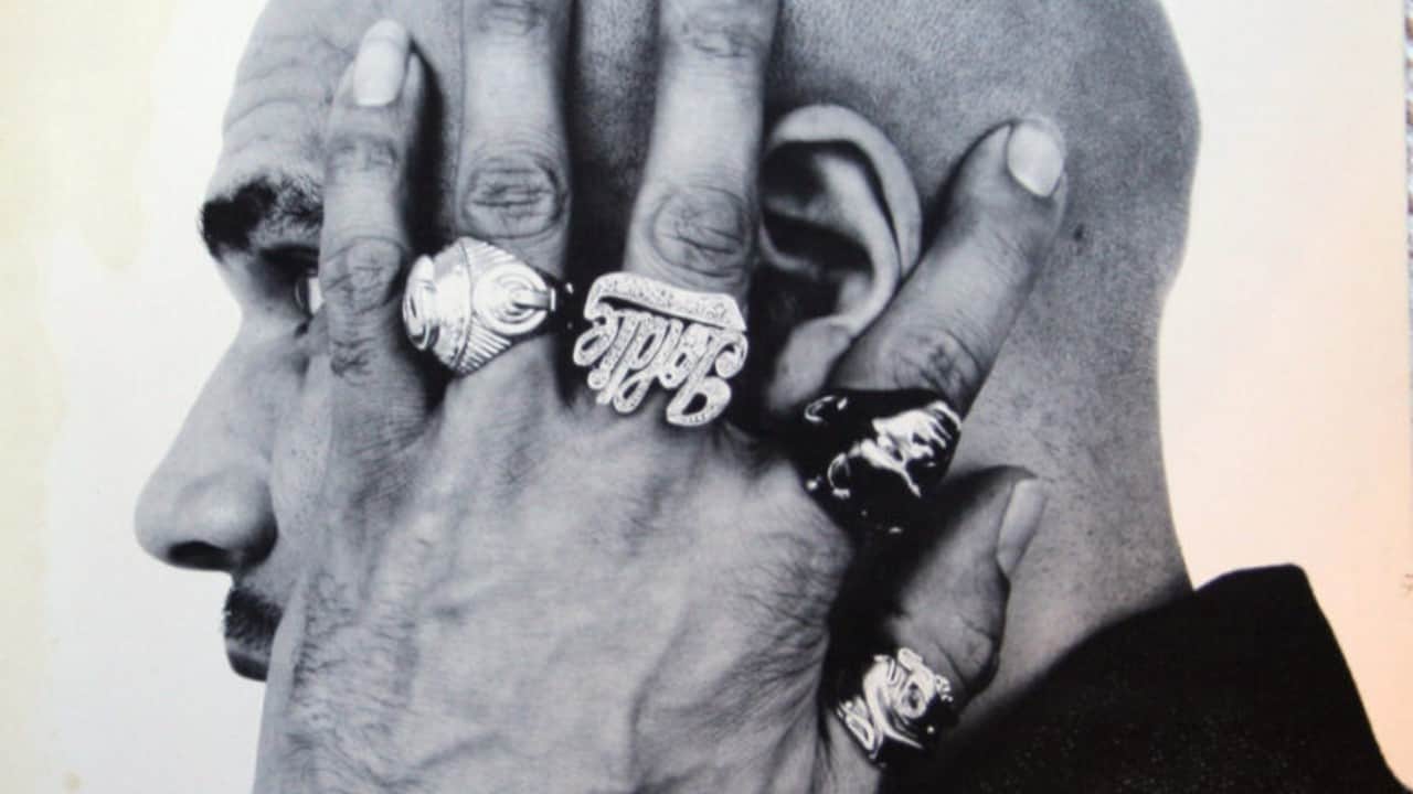 A black-and-white photo of Goldie (real name Clifford Joseph Price MBE) from a profile angle covering his face with rings on his fingers.